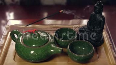 佛像在木茶板上用绿茶壶和杯子。 燃烧着的香枝散发出的香气